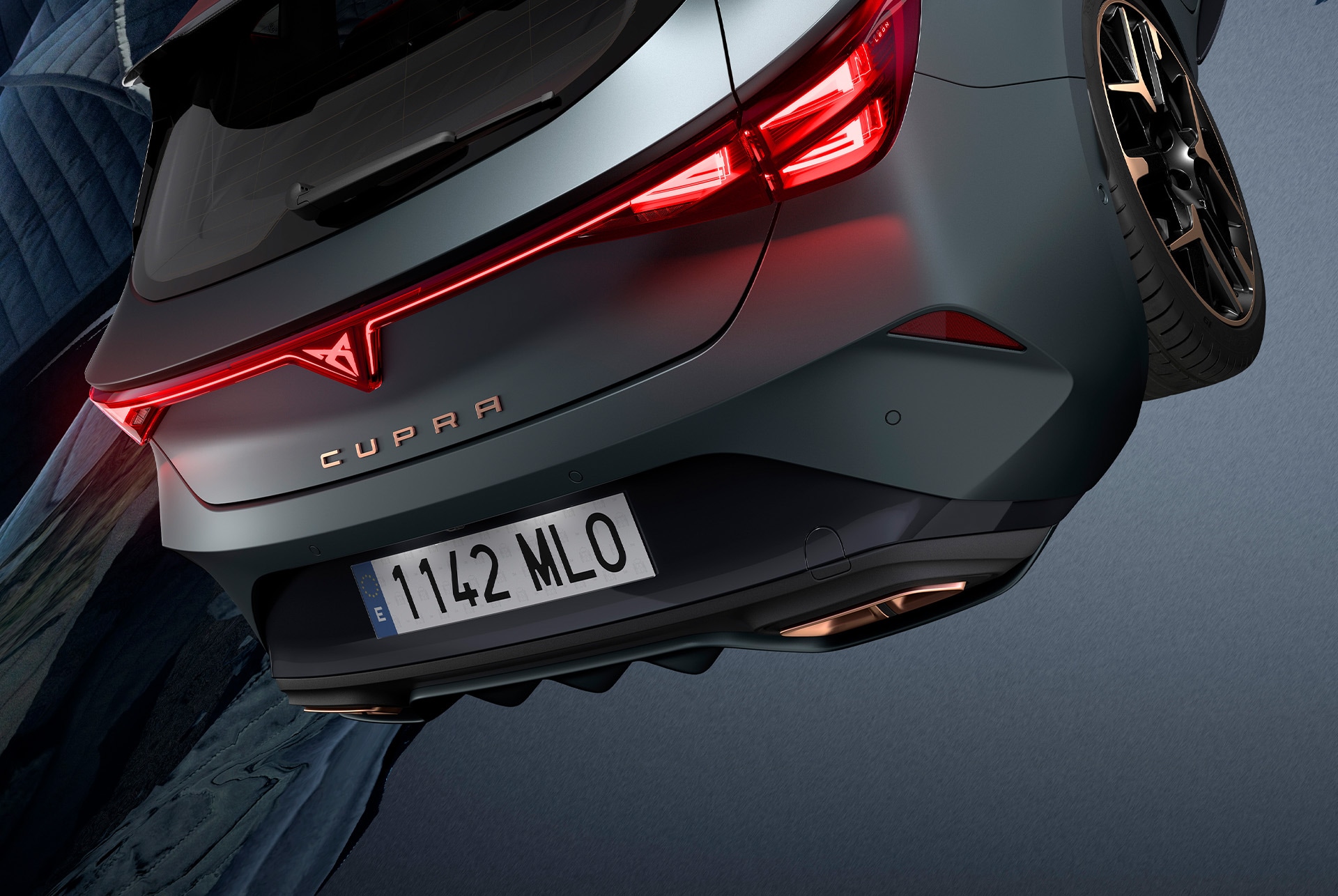 CUPRA Leon in matte grey is parked on a rooftop at night. A person is performing a dynamic jump in front of the car, adding a sense of thrill to the scene. The city skyline is visible in the background, with its lights creating a vibrant glow. The car is illuminated by red lights, highlighting its sporty design and aggressive front end