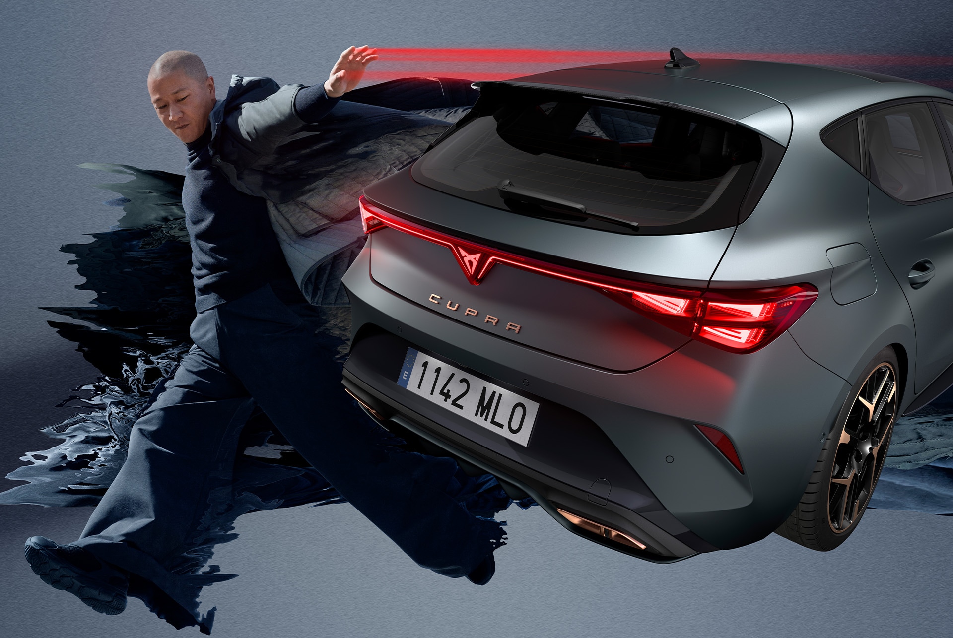 A sleek, matte grey CUPRA Leon is parked on a city street at night. A person is performing a dynamic jump in front of the car, adding a sense of energy and movement to the scene. The car is illuminated by red lights, highlighting its sporty design and aggressive front end.